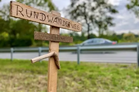 Hier geht es zum berühmten "Sauerborn": Dieses Holzschild am Ortsrand von Lindenholzhausen ist Ausdruck des Bürgerprotests, denn eine neue Leitplanke versperrt Spaziergängern an dieser Stelle die Überquerung der Bundesstraße 8 (im Hintergrund), die den Rundweg regelmäßig nutzen.